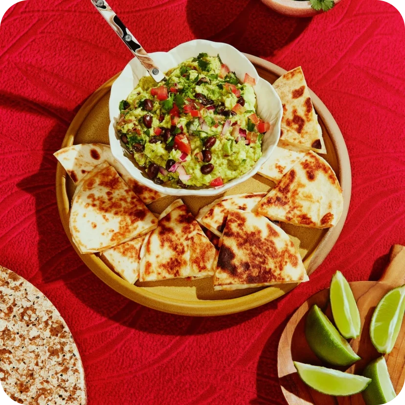 a dinner table with a bright red table cloth and a plate of Quesadillas surrounding a bowl of Guacamole